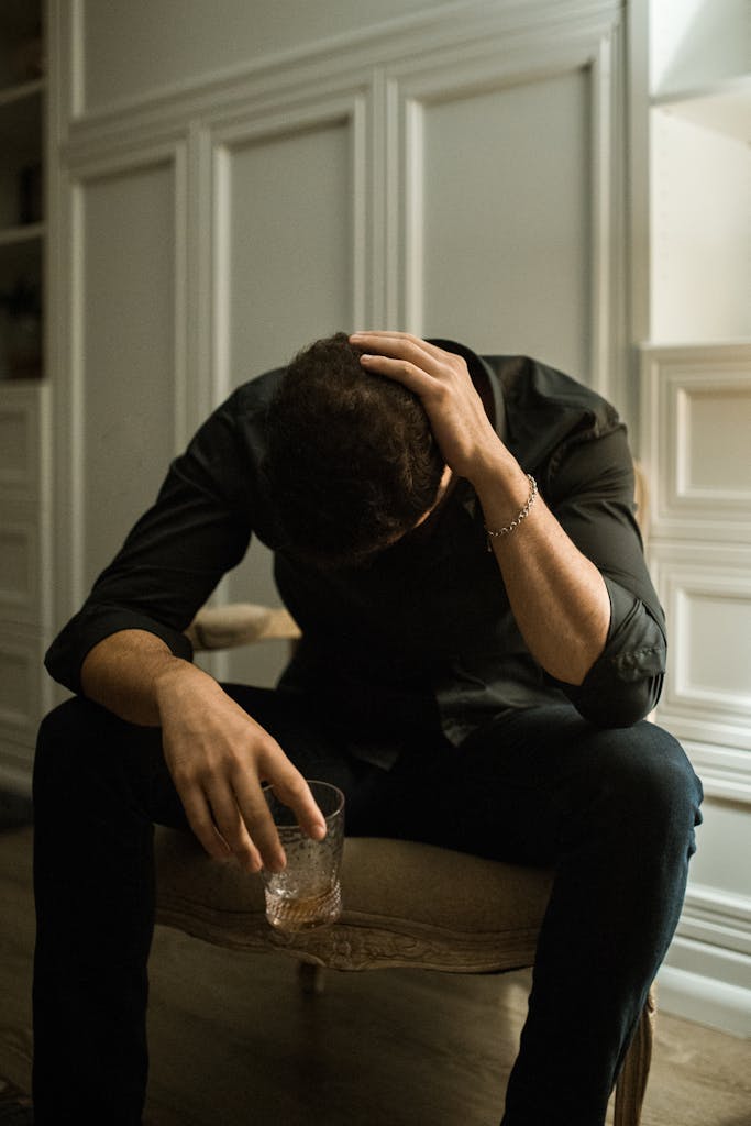 Man in distress seated indoors, head in hand, holding a drink, reflecting solitude.